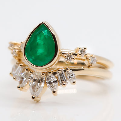 A pear-shaped emerald set in a gold bezel ring, adorned with offset small diamonds on the band. Stacked with an intricate crown-like diamond band. Displayed on a white background.
