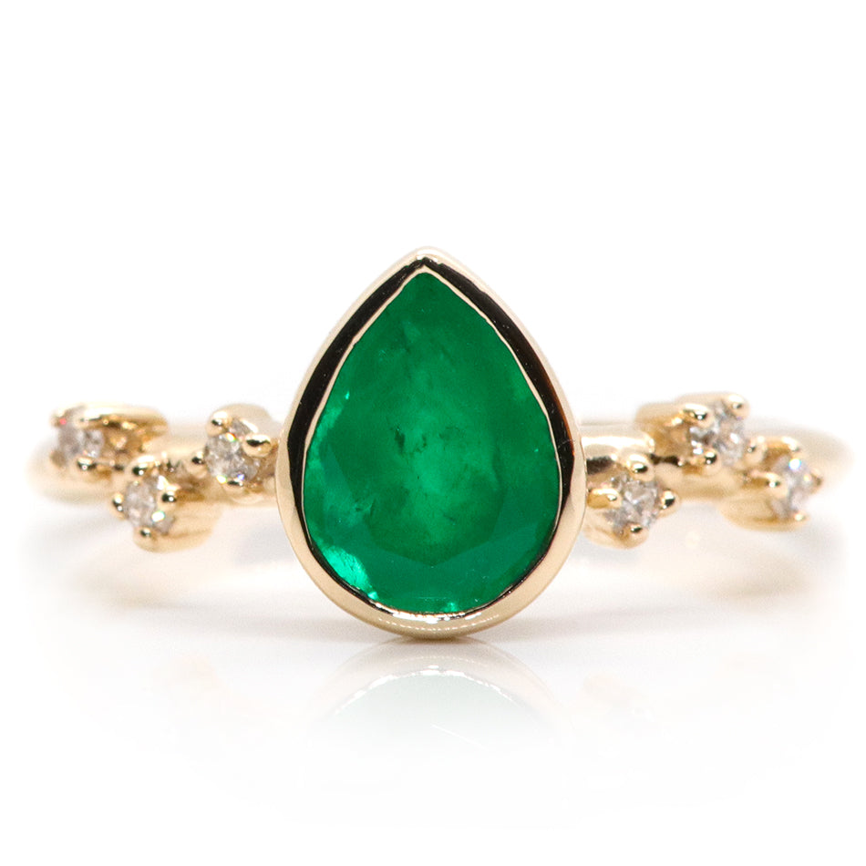 A pear-shaped emerald set in a gold bezel ring, adorned with offset small diamonds on the band. Displayed on a white background 
