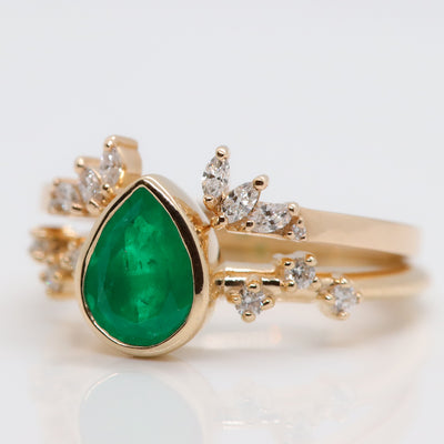 A pear-shaped emerald set in a gold bezel ring, adorned with offset small diamonds on the band. Stacked with an intricate open diamond band. Displayed on a white background.