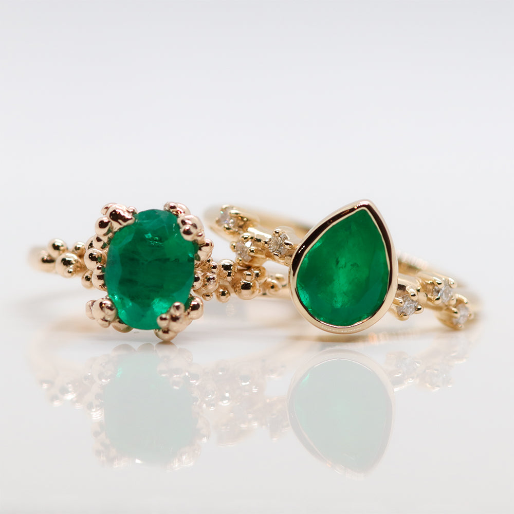 Two gold rings with vibrant green emerald gemstones; the left ring features an oval cut, and the right has a pear-shaped design. The pear-shaped ring is accented with small diamonds, exuding elegance and sophistication.