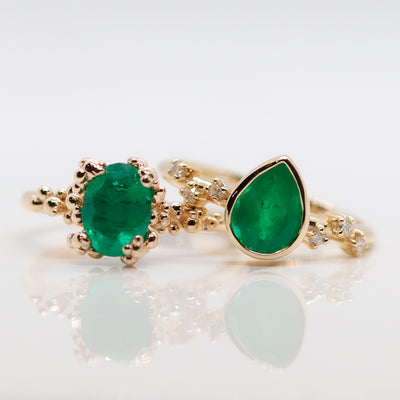 Two gold rings with vibrant green emerald gemstones; the left ring features an oval cut, and the right has a pear-shaped design. The pear-shaped ring is accented with small diamonds, exuding elegance and sophistication.