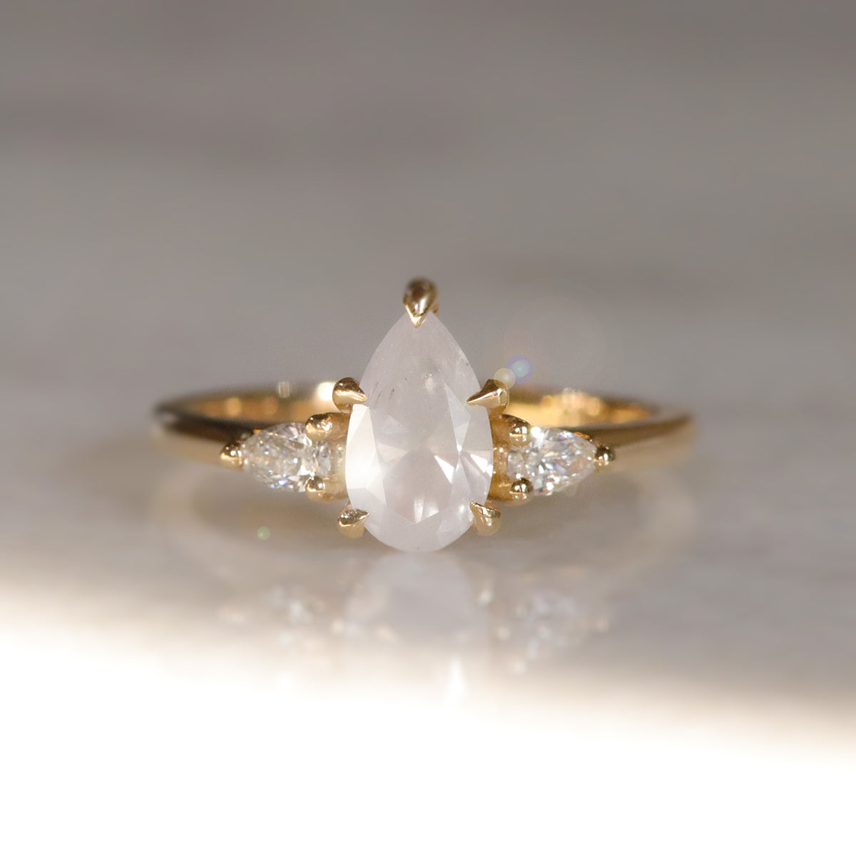 A three stone ring with a central fancy white pear-shaped diamond set in gold , flanked by two smaller pear diamonds, on a soft, light, blurred background.