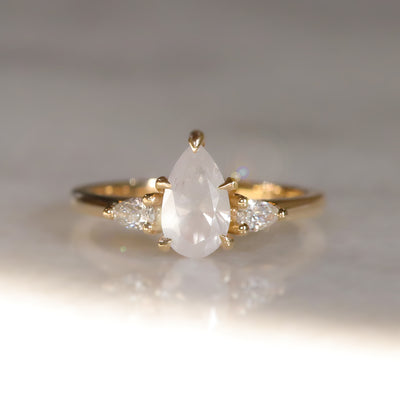 A three stone ring with a central fancy white pear-shaped diamond set in gold , flanked by two smaller pear diamonds, on a soft, light, blurred background.