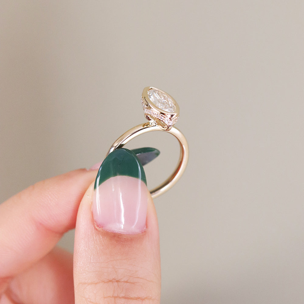 A side profile view of an elongated marquise-cut diamond ring bezel set with a hidden diamond halo and a polished gold band, shown held between fingers. 