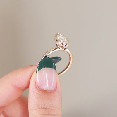A side profile view of an elongated marquise-cut diamond ring bezel set with a hidden diamond halo and a polished gold band, shown held between fingers. 