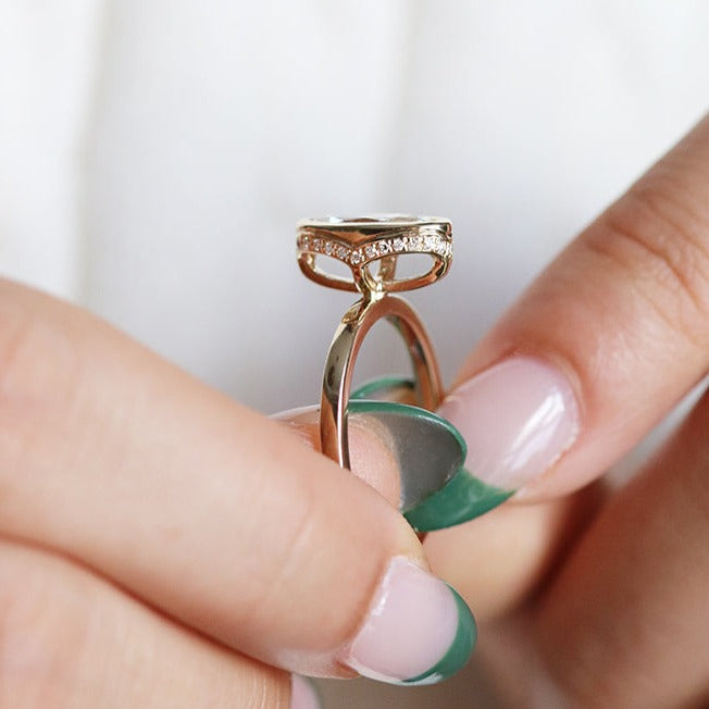 A side profile view of an elongated marquise-cut diamond ring bezel set with a hidden diamond halo and a polished gold band, shown held between fingers. 