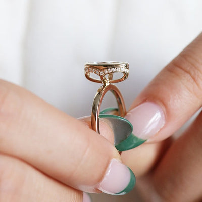 A side profile view of an elongated marquise-cut diamond ring bezel set with a hidden diamond halo and a polished gold band, shown held between fingers. 