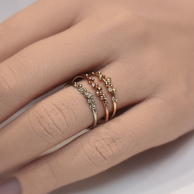 Three delicate rings in different types of gold including rose gold, yellow gold and white gold, featuring unique organic designs with small bead-like accents and sparkling diamonds, stacked on a finger.