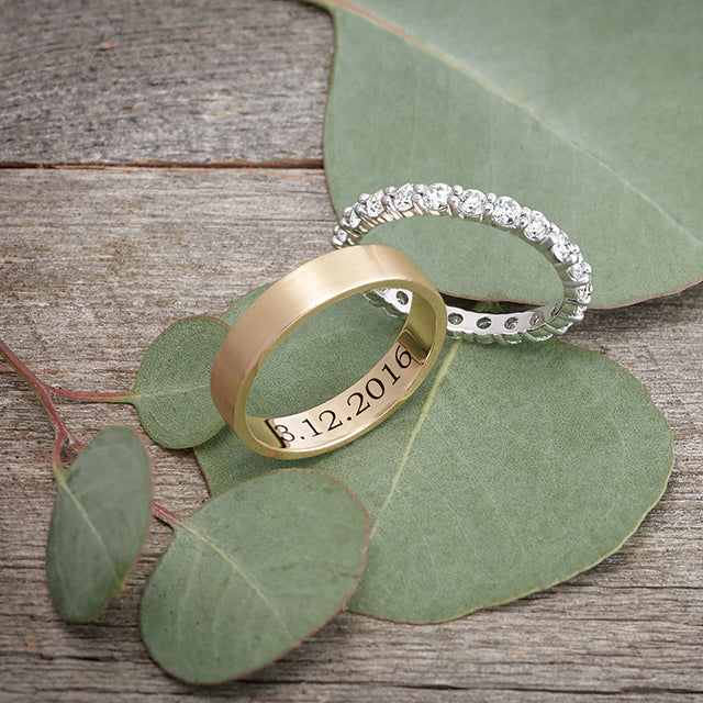 A simple flat edge gold wedding band engraved with a special date, resting next to a diamond white gold eternity band on a background of elegant foliage.