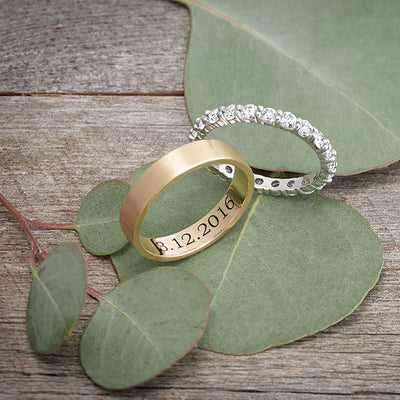 A simple flat edge gold wedding band engraved with a special date, resting next to a diamond white gold eternity band on a background of elegant foliage.
