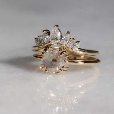 Peaked gold ring sparkles with 7 diamonds in oval, pear, marquise and rectangele shapes to form a crown appearance. Resting on round solitaire ring with a rosecut diamond on a white background