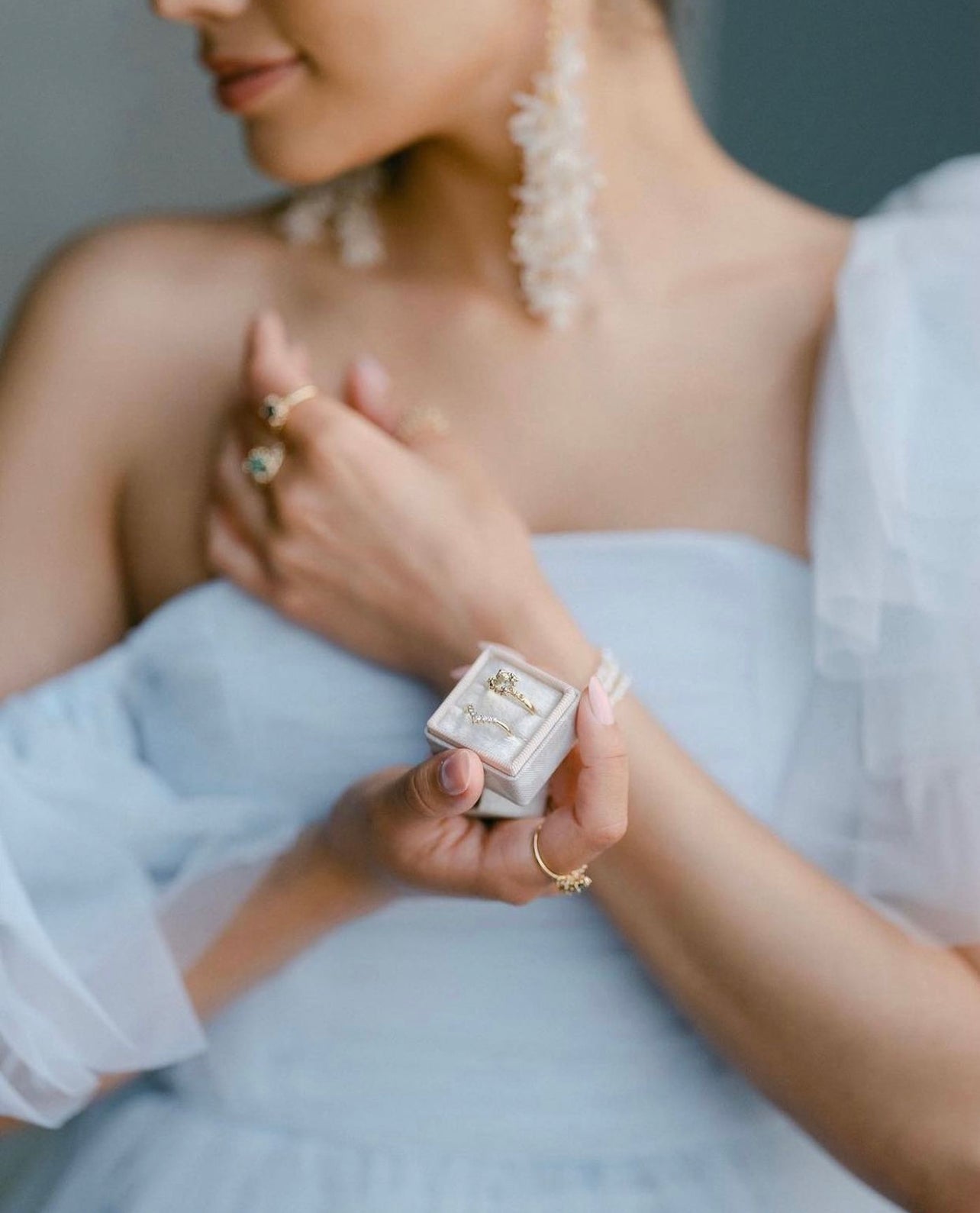 A woman in pretty bridal wear holds a small square velvet ring box, containing a green sapphire engagement ring and diamond wedding band. She wears dramatic pearl earrings and other stacking rings on her fingers.