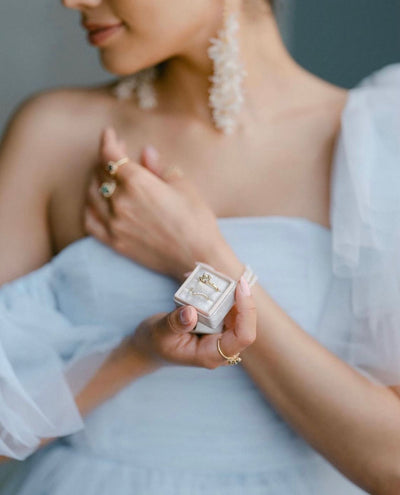 A woman in pretty bridal wear holds a small square velvet ring box, containing a green sapphire engagement ring and diamond wedding band. She wears dramatic pearl earrings and other stacking rings on her fingers.