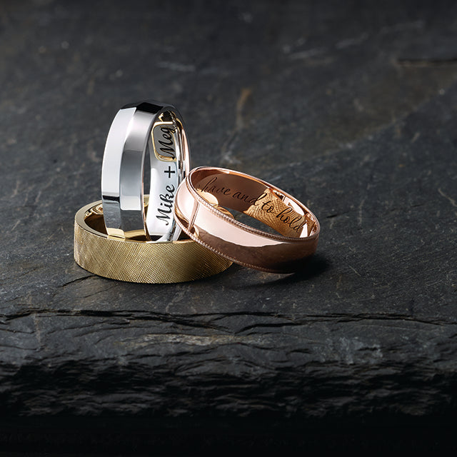 Three elegant wedding rings displayed on a slate surface: a silver band, a gold band, and a rose gold band engraved with special messages.